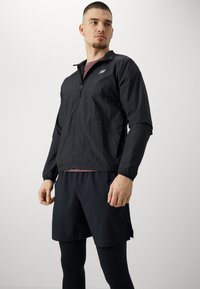 Veste légère noire avec fermeture éclair sur le devant, manches longues et poignets élastiques, associée à un short noir et des leggings ; détails de marque minimalistes.