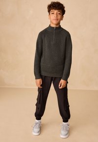 Maglione verde scuro lavorato a maglia con collo alto, abbinato a pantaloni cargo neri con tasche laterali e polsini elasticizzati, indossato con scarpe da ginnastica bianche.