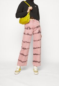 Persona che indossa jeans ampi a vita alta rosa tie-dye, giacca corta nera, sneakers giallo-bianche e tiene una borsa a tracolla gialla.