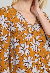 Blouse moutarde avec un col en V, ornée d'un imprimé floral blanc avec des contours marron. Tissu doux avec une coupe décontractée et un design fluide.