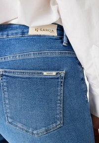 Nahaufnahme einer blauen Denim-Jeans mit Gesäßtasche und Garcia-Label, getragen mit einem eingeführten weißen Hemd.