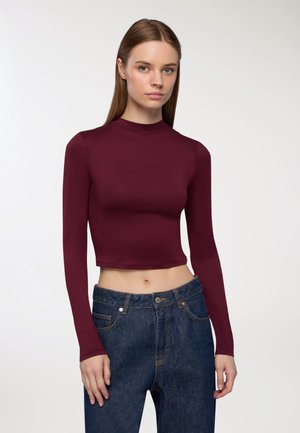 Top corto a maniche lunghe di colore bordeaux con collo alto, realizzato in un tessuto liscio. Abbinato a jeans in denim scuro a vita alta.
