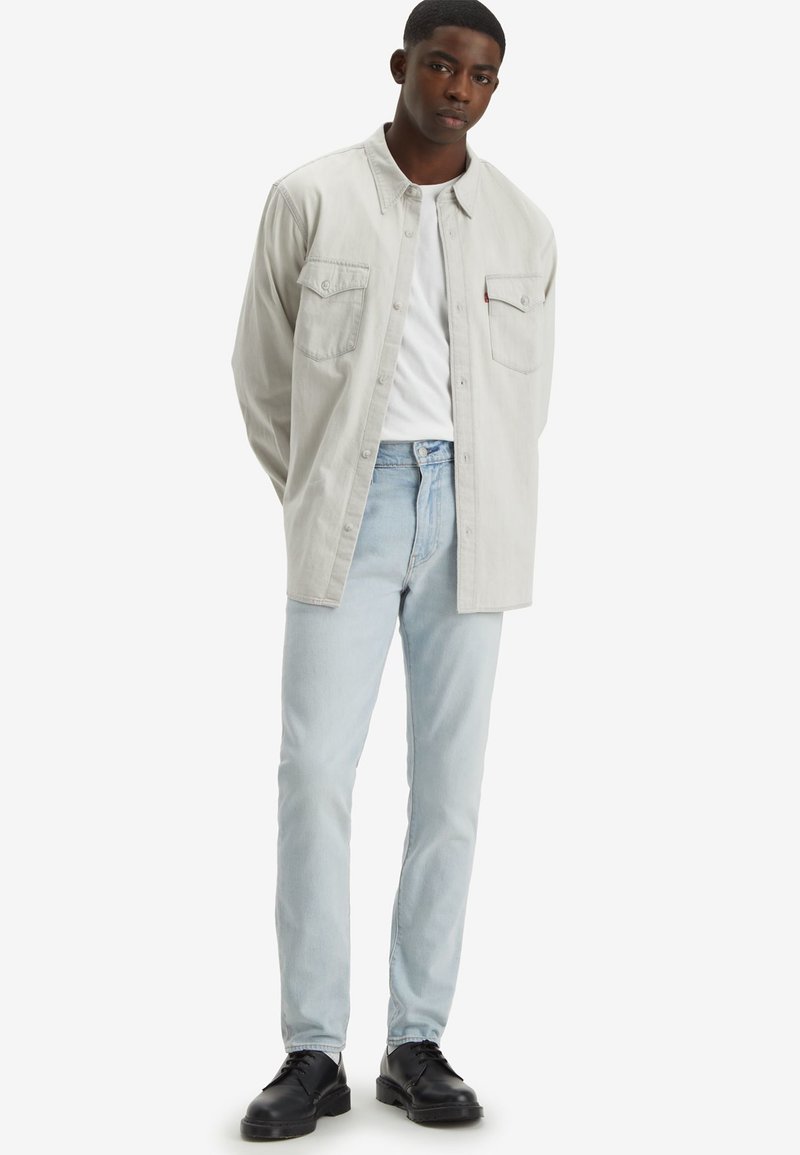Camisa de botones gris claro sobre una camiseta blanca, combinada con jeans azul claro y zapatos negros. Presenta dos bolsillos en el pecho.