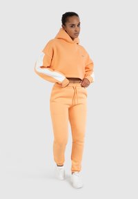 Geknipt oranje hoodie met witte accenten op de mouwen, gecombineerd met bijpassende oranje joggingsbroeken en witte sneakers. Zachte katoenen mengsel.