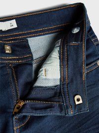 Mørkeblå denimjeans med gullsøm, en synlig glidelås og en liten lomme. Livsermerket har en knapp og et stoffmerke.