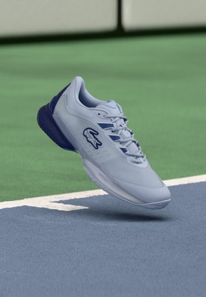 Zapatilla deportiva azul claro con una parte superior lisa, malla texturizada y suela azul oscuro. Cuenta con un logotipo en el lateral y detalles en contraste.