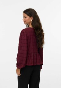 Blouse texturée bordeaux avec un ourlet peplum froncé et des manches ballons, portée avec un pantalon noir. Cheveux coiffés en vagues lâches.