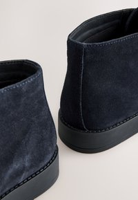 Marineblaue Wildleder-Stiefeletten mit glatter Textur, abgerundetem Design und schwarzen Gummisohlen. Gestickte Details entlang der Sohle.