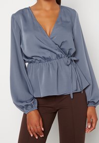 Kvinna i en dammig blå omslagsblus med långa puffärmar och bruna åtsittande byxor, stående med händerna avslappnat vid sidorna.