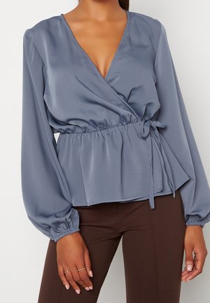 Frau in einer staubblauen Wickelbluse mit langen Puffärmeln und braunen, taillierten Hosen, die mit entspannten Händen an den Seiten steht.