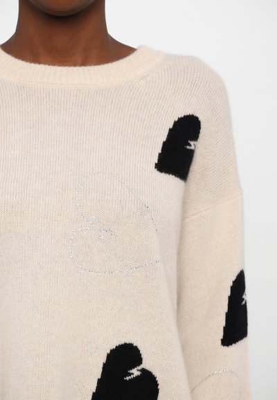 Beżowy sweter dziany z czarnym wzorem w serca i srebrnym detalem serca. Posiada szeroki dekolt i luźny krój. Miękka, teksturowana tkanina.