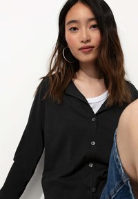 Gilet à boutons noirs avec un col, présentant une texture douce, porté sur un haut blanc à plusieurs couches, avec des cheveux foncés de longueur moyenne visibles.