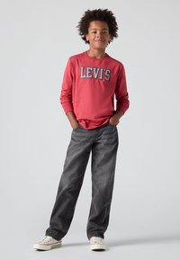 Camisa roja de manga larga con el logo "LEVI'S", combinada con jeans grises y zapatillas blancas. La camisa tiene puños ajustados y un corte relajado.