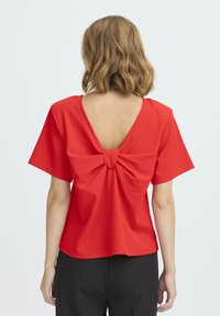 Top rojo de manga corta hecho de tela lisa, con un escote en V profundo en la espalda y un detalle de nudo en el centro. Diseño liso, corte relajado.