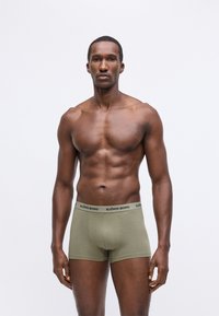 Fekete bőrű, izmos férfi egyenesen áll, olíva zöld boxer alsót visel, amelynek derékrészén a "Björn Borg" logó található, semleges szürke háttérrel.