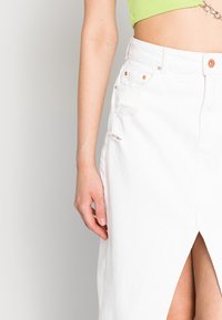 Jupe en denim blanc avec une fente haute, présentant des détails déchirés et un matériel de boutons en ton cuivre. Texture lisse et design mi-genoux.