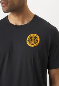 Schwarzes T-Shirt aus Baumwolle mit rundem Kragen und kurzen Ärmeln. Auffälliges oranges rundes Logo mit Text im linken Brustbereich.