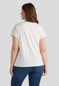 Camiseta blanca de manga corta con detalles de ojales en las mangas, combinada con pantalones vaqueros azules, vista desde atrás. Textura suave.