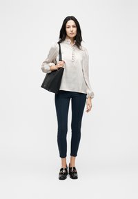 Γυναίκα που φοράει μπεζ ριγέ μπλούζα, στενά σκούρα μπλε παντελόνια, μαύρα μοκασίνια με χρυσή λεπτομέρεια και κρατάει μια μεγάλη μαύρη τσάντα ώμου.