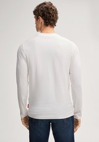 Weißes Langarmshirt aus leichtem Stoff mit Rundhalsausschnitt und dezentem Markenlogo auf der Rückseite, kombiniert mit dunkler Denim-Jeans.