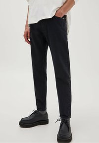 Pantalones de mezclilla azul oscuro con un ajuste slim, textura sutil, dos bolsillos frontales y un diseño clásico de cinco bolsillos, combinados con zapatos negros.