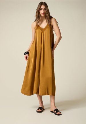 Donna con capelli mossi sciolti che indossa un vestito midi a sottoveste color senape, sandali neri a ciabatta e bracciali neri grossi al polso sinistro.