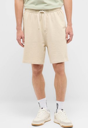 Man wearing beige drawstring shorts, white branded socks, and white sneakers standing against a plain background.