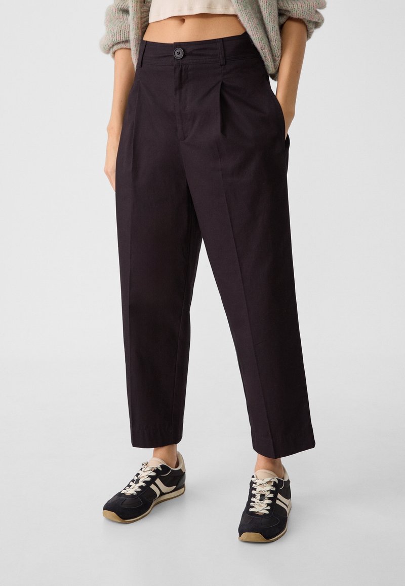 Femme portant un pantalon noir taille haute fuselé avec des plis devant et des baskets décontractées noires et blanches, debout avec une main dans la poche.