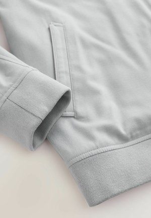 Mâneca unui hanorac gri deschis cu manșetă ribbed și detaliu de buzunar pe fundal de material moale.