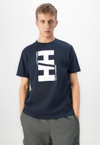 Junger Mann mit kurzen Haaren, der ein marineblaues Helly Hansen T-Shirt und graue Hosen trägt, steht mit den Händen in den Taschen vor einem weißen Hintergrund.