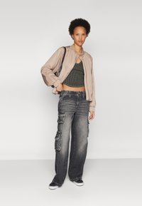 Veste bomber beige clair avec des poignets élastiques, haut court côtelé vert olive, jean cargo ample noir avec poches latérales et baskets noires.