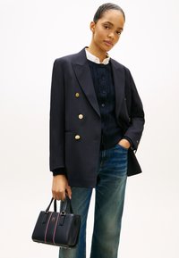 Γυναίκα που φοράει μαύρο σακάκι διπλής σειράς με χρυσά κουμπιά, μπλεζιά καλωδίωσης ναυτικού χρώματος, μπλε τζιν και κρατάει μαύρη τσάντα χειρός.