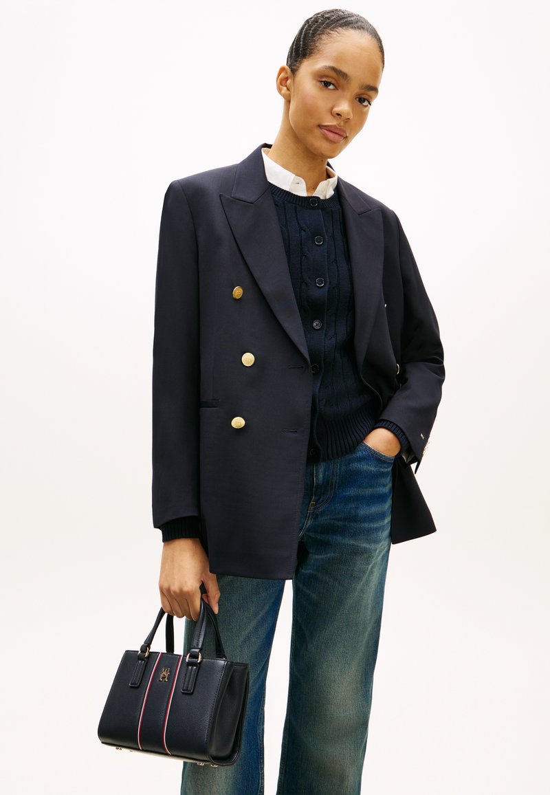 Γυναίκα που φοράει μαύρο σακάκι διπλής σειράς με χρυσά κουμπιά, μπλεζιά καλ ωδίωσης ναυτικού χρώματος, μπλε τζιν και κρατάει μαύρη τσάντα χειρός.