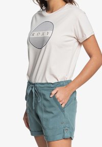Světle růžové tričko s námořnickou grafikou, která zobrazuje pruhovaný ovál a text "ROXY", spárované s tyrkysovými šortkami na stahování.