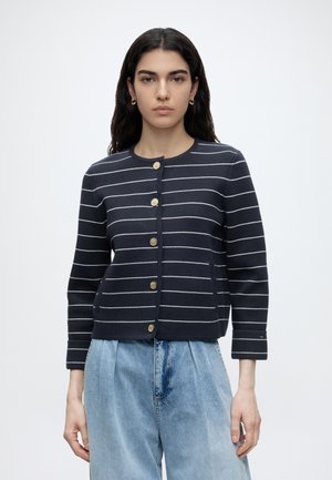 Donna con capelli scuri indossa un cardigan blu navy con strisce orizzontali bianche e bottoni dorati, abbinato a jeans a vita alta azzurri chiari.