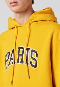 Sweat à capuche jaune avec "PARIS" imprimé en noir et blanc. Doté d'une capuche à cordon de serrage et d'une texture douce. Gros plan sur l'encolure et la zone de la poitrine.