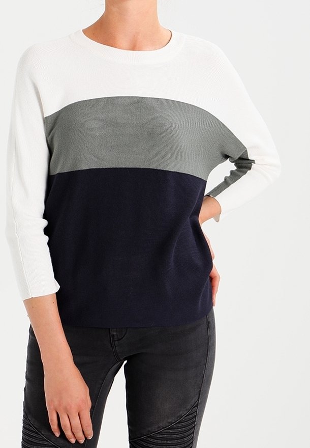 Personne portant un pull à blocs de couleur avec des sections blanches, grises et bleu marine, associées à un pantalon noir texturé, debout devant un fond uni.