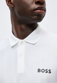 Gros plan d’un homme portant un polo blanc à col avec le logo « BOSS » sur la poitrine, regardant sur le côté sur un fond uni.