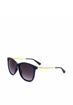 Occhiali da sole neri a forma di occhio di gatto con lenti sfumate scure e aste dorate testurizzate con un piccolo logo Ted Baker ai lati.
