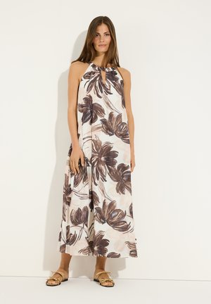 Vrouw draagt een mouwloze, enkellange witte jurk met grote donkere bloemprint en beige platte sandalen, staand tegen een witte muur.