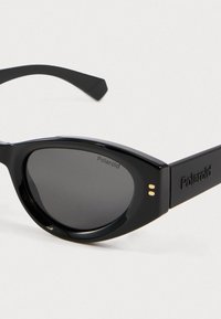 Μαύρα γυαλιά ηλίου cat-eye με σκούρους φακούς, γυαλιστερό πλαστικό σκελετό και ανάγλυφο λογότυπο "Polaroid" στους βραχίονες. Στενοί βραχίονες.