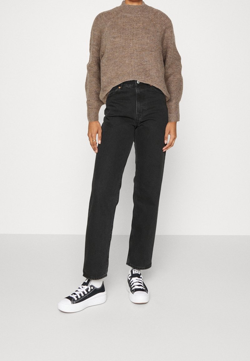 Personne portant un pull en maille marron, un jean noir taille haute, des chaussettes blanches et des baskets plateformes noires et blanches, debout sur un fond uni.