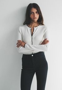 Top alb cu mâneci lungi, cu dungi și tiv negru, cu închidere cu nasturi; asortat cu blugi înalți, întunecați. Modelul pozează cu brațele încrucișate.