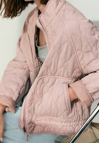 Veste matelassée rose clair avec fermeture éclair à l'avant, col montant, poches latérales et ourlet élastique, fabriquée en tissu doux à motif texturé.