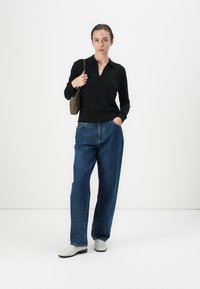 Zwarte gebreide polo met een v-hals, gecombineerd met brede blauwe denim jeans. Witte schoenen en een kleine groene handtas maken de outfit compleet.