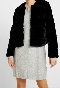 Kvinde iført en sort faux fur jakke over en lys grå strikket kjole, stående med hænderne afslappet ved sin side.