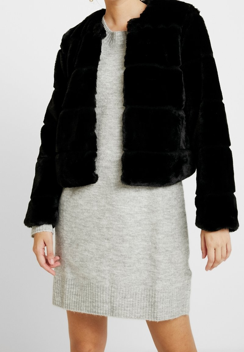 Kvinde iført en sort faux fur jakke over en lys grå strikket kjole, stående med hænderne afslappet ved sin side.