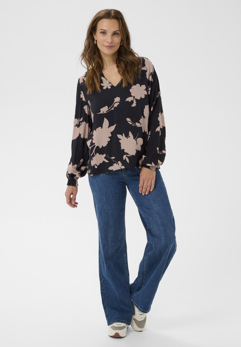 Zwarte blouse met groot beige bloemenpatroon, V-hals, lange mouwen met gathered manchetten, gecombineerd met blauwe flared jeans en witte sneakers.