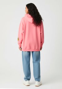 Sudadera rosa de gran tamaño con capucha, que presenta un gráfico de sol en la manga izquierda. Combinada con pantalones azules claros con estampado y zapatillas blancas.