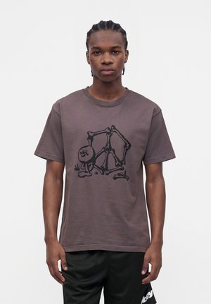 Jeune homme aux cheveux tressés portant un t-shirt marron présentant un dessin squelettique abstrait noir et un short de sport noir.